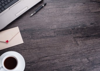 Bureau de couleur sombre en bois avec un ordianteur portable en haut à gauche, un café un stylo et une enveloppe avec un cœur rouge.