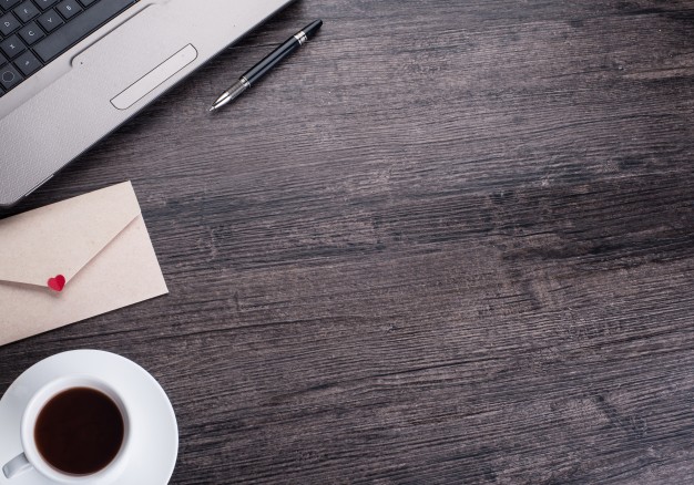 Bureau de couleur sombre en bois avec un ordianteur portable en haut à gauche, un café un stylo et une enveloppe avec un cœur rouge.